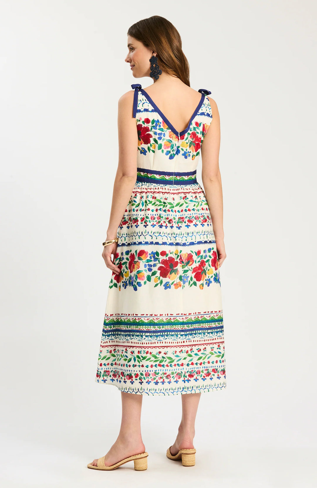 Back of Tyler Boe Chelsey Cotton Summer Soiree Dress in ivory with multicolor painterly floral print and navy grosgrain trim, shown front on-model, square V-neckline with shoulder-tie straps, fitted bodice, full midi skirt with bordered print bands, styled with raffia slides.