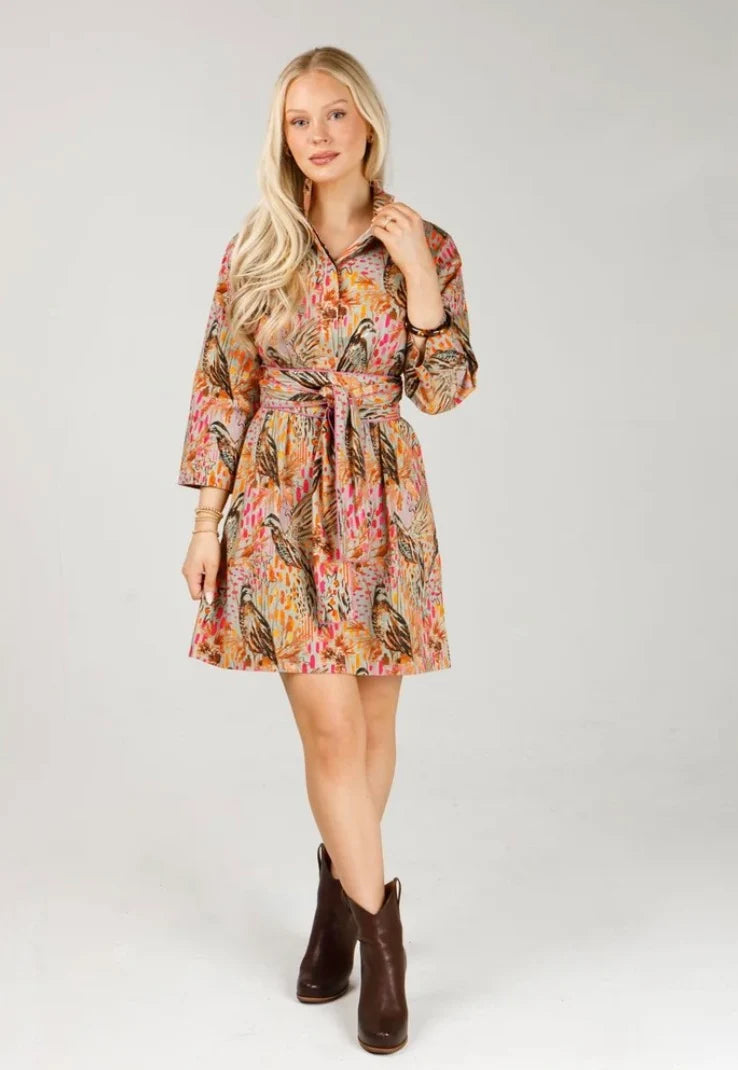 Holly Shae Arden Dress in Wildwood Wing, full-length front view on model against a light gray backdrop, showing multicolor game bird and feather print, stand collar with button placket, three-quarter sleeves, self-tie sash at the waist, and above-knee hem with brown ankle boots.