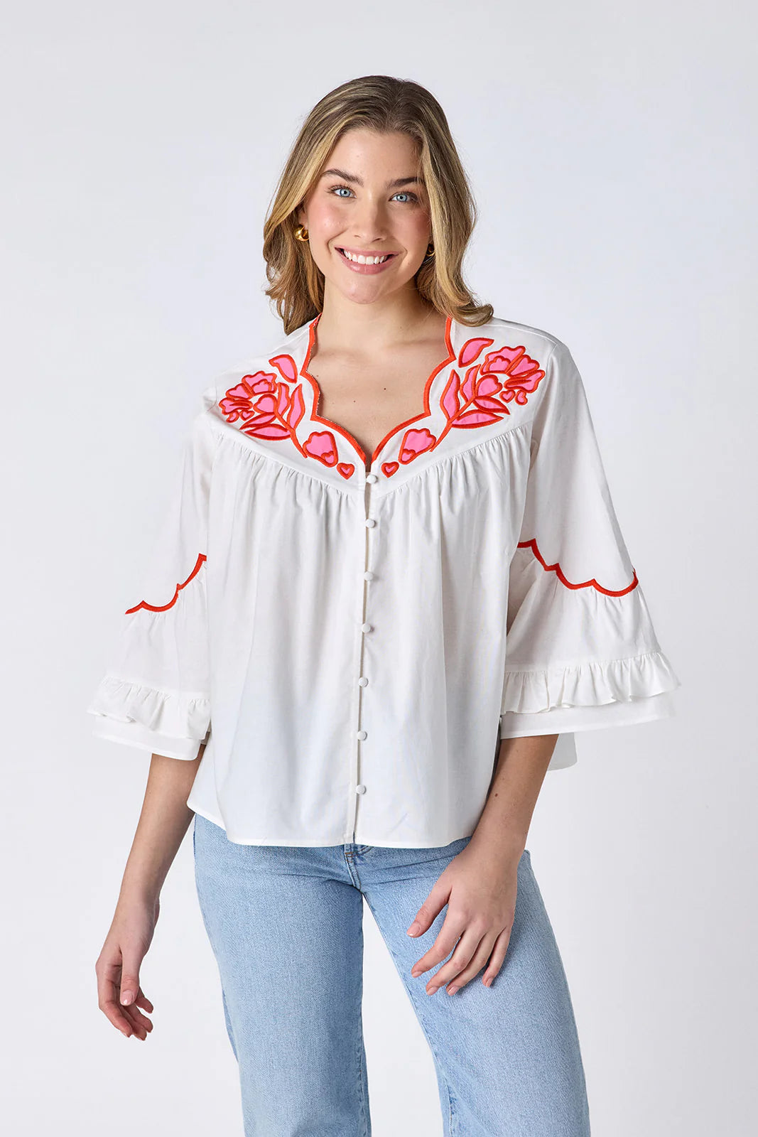 Model wearing Crosby Ity Blouse in ivory, front view, showing coral and pink floral embroidered yoke, scalloped V-neckline, and ruffle-cuffed bell sleeves, styled with light-wash straight-leg jeans and gold earrings, on grey background.