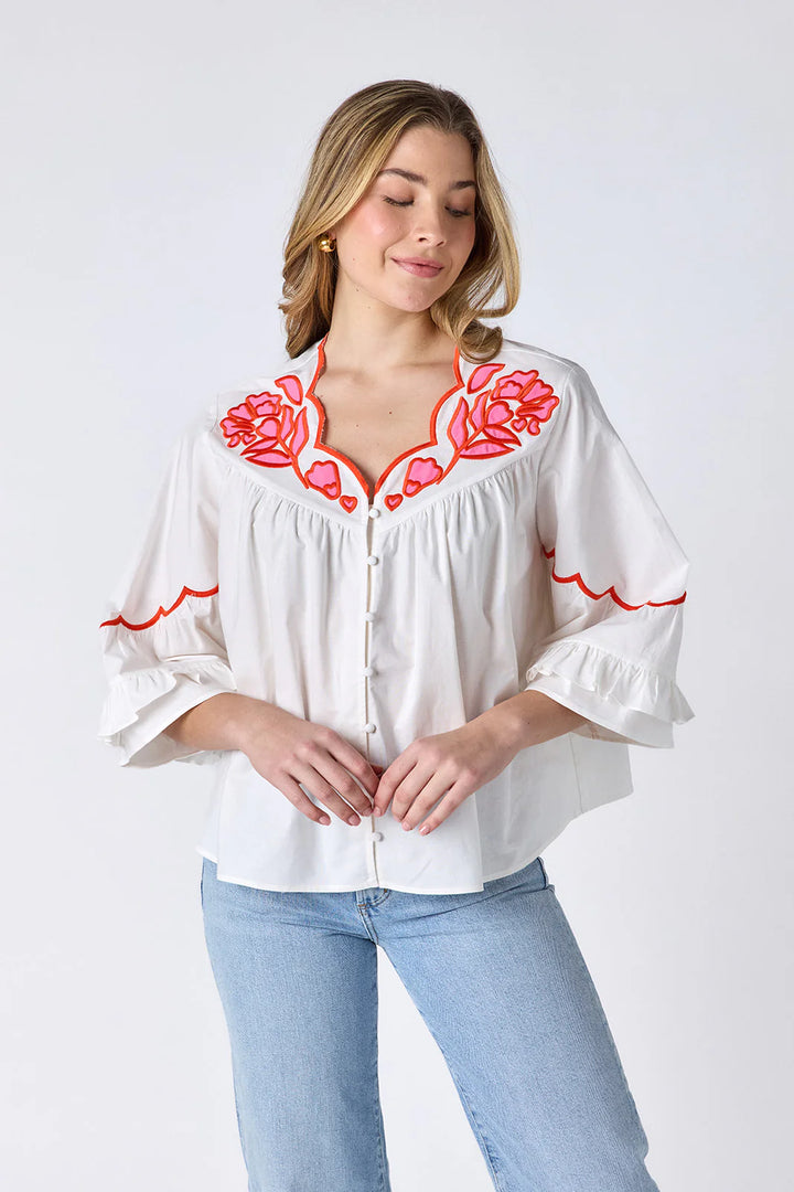 Model wearing Crosby Ity Blouse in ivory, front view, showing coral and pink floral embroidered yoke, scalloped V-neckline, and ruffle-cuffed bell sleeves, styled with light-wash straight-leg jeans and gold earrings, on grey background.