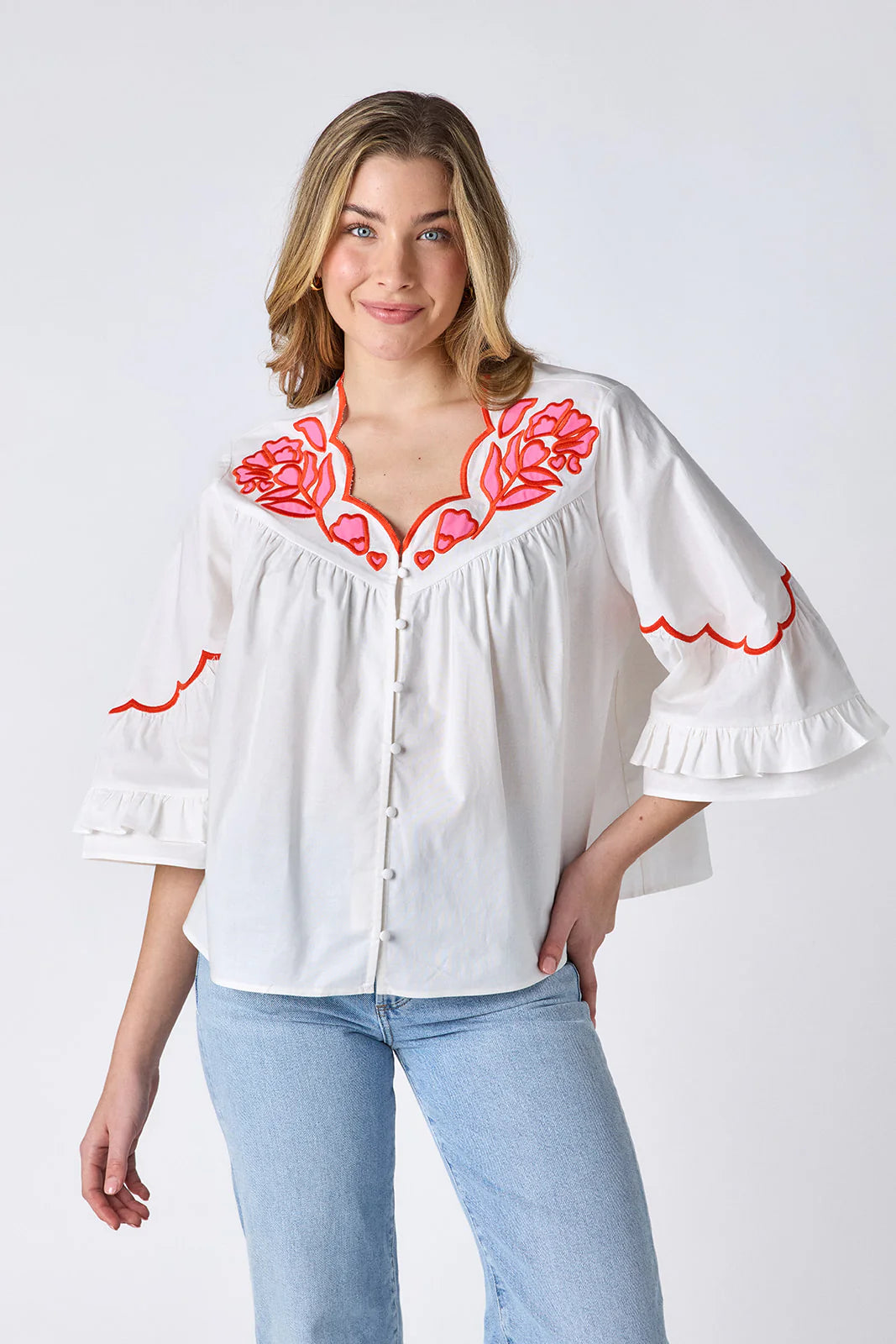 Model wearing Crosby Ity Blouse in ivory, front view, showing coral and pink floral embroidered yoke, scalloped V-neckline, and ruffle-cuffed bell sleeves, styled with light-wash straight-leg jeans and gold earrings, on grey background.