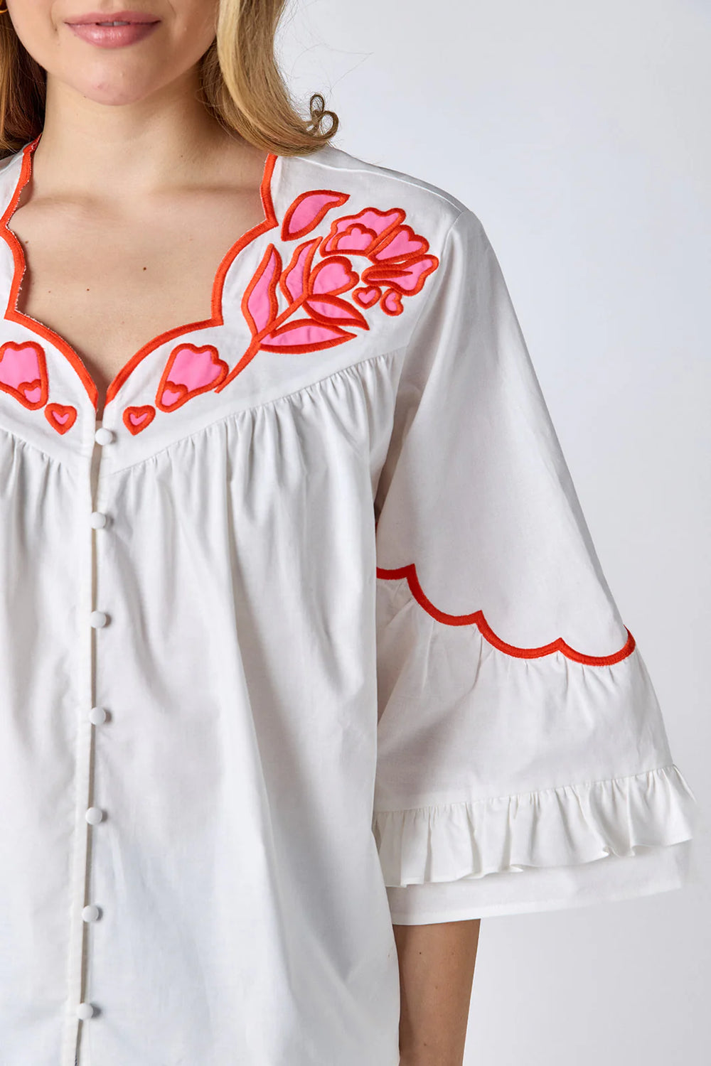 Model wearing Crosby Ity Blouse in ivory, front view, showing coral and pink floral embroidered yoke, scalloped V-neckline, and ruffle-cuffed bell sleeves, styled with light-wash straight-leg jeans and gold earrings, on grey background.