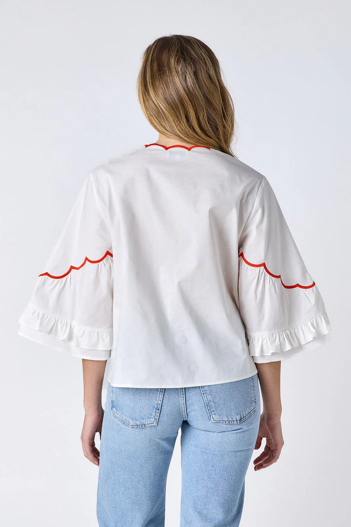 Model wearing Crosby Ity Blouse in ivory, front view, showing coral and pink floral embroidered yoke, scalloped V-neckline, and ruffle-cuffed bell sleeves, styled with light-wash straight-leg jeans and gold earrings, on grey background.
