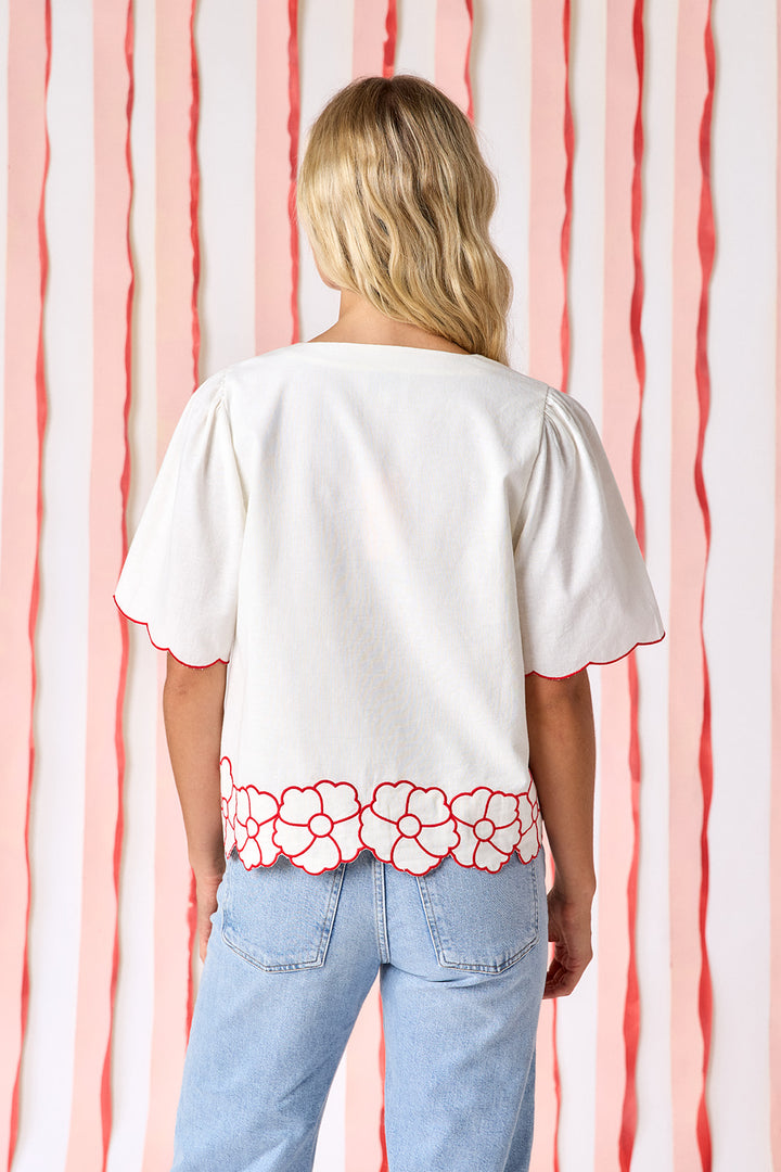 Model wearing Crosby Jane Short Sleeve Top in ivory styled with light-wash straight-leg jeans. Top features red contrast piping at neckline and scalloped sleeves, floral appliqué hem border, and fabric-covered button-front closure - back