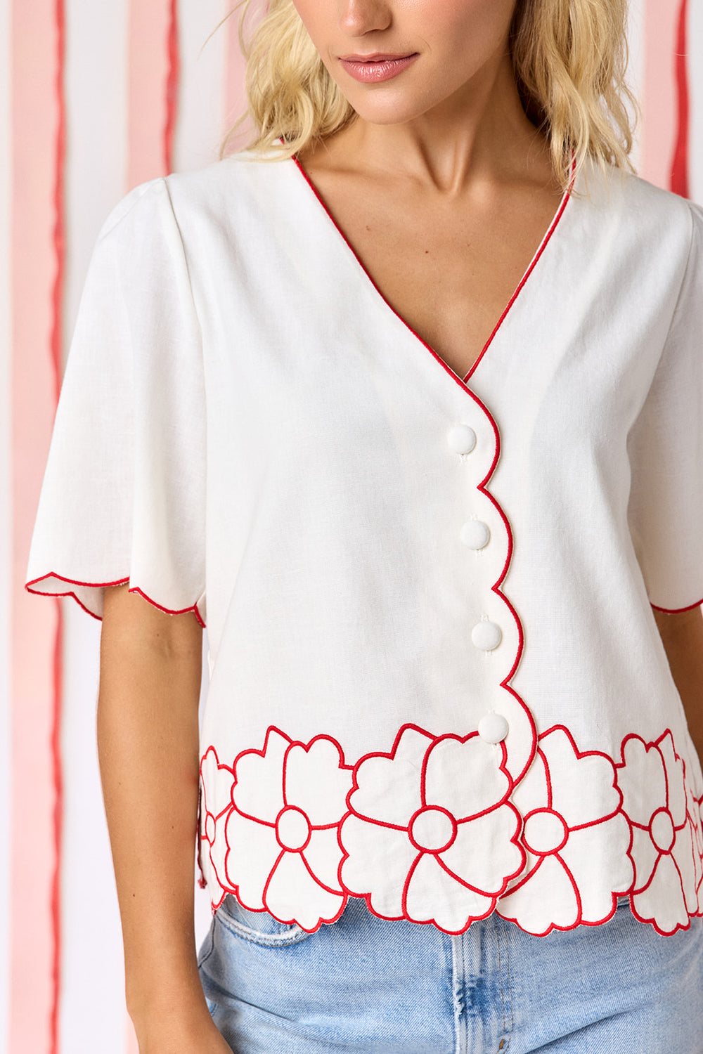 Model wearing Crosby Jane Short Sleeve Top in ivory styled with light-wash straight-leg jeans. Top features red contrast piping at neckline and scalloped sleeves, floral appliqué hem border, and fabric-covered button-front closure - closeup