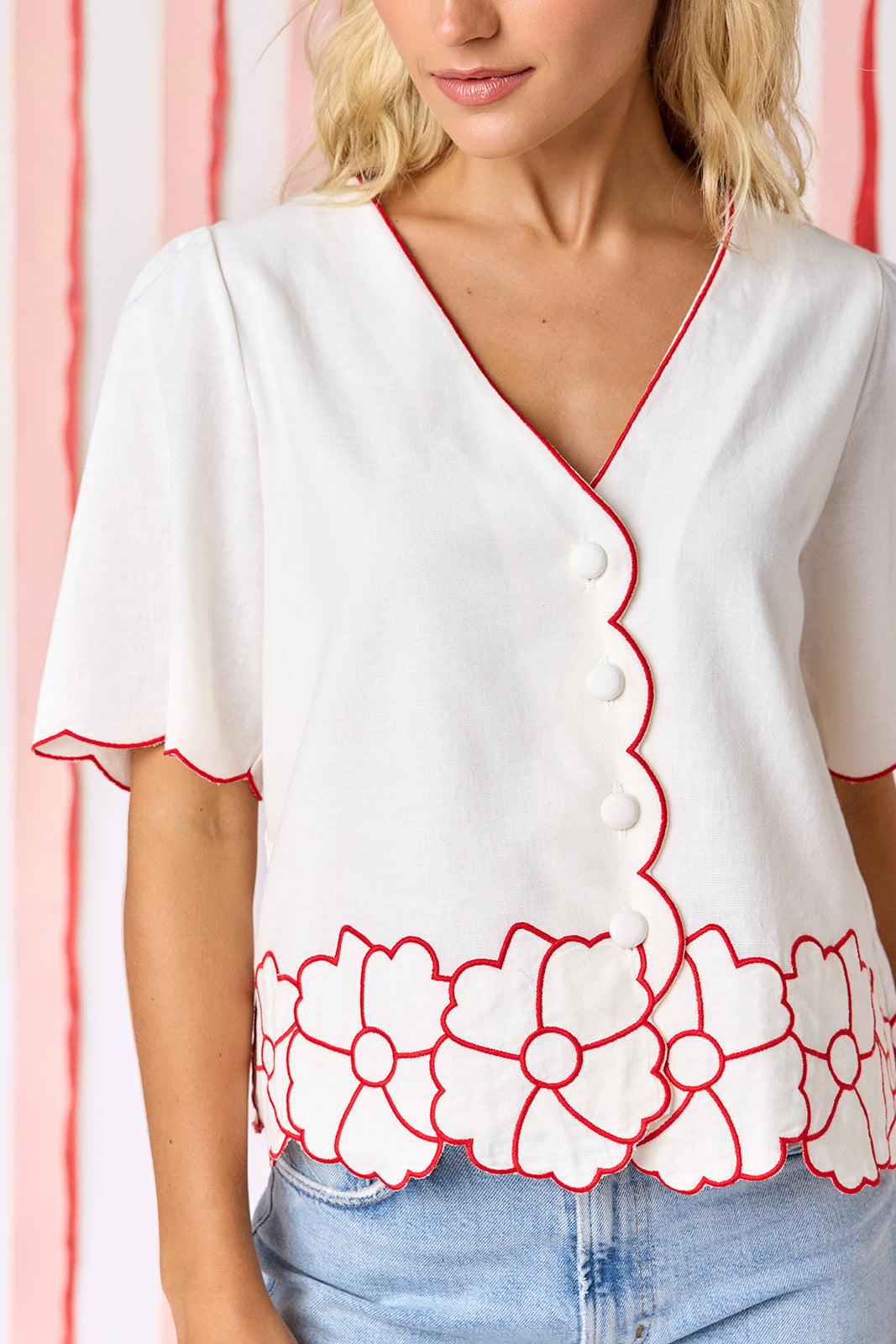 Model wearing Crosby Jane Short Sleeve Top in ivory styled with light-wash straight-leg jeans. Top features red contrast piping at neckline and scalloped sleeves, floral appliqué hem border, and fabric-covered button-front closure - closeup