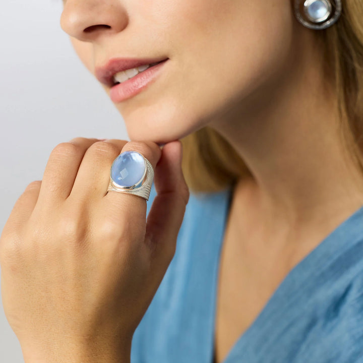 Julie Vos Verona Statement Ring in silver plate — wide chevron-etched band with large oval faceted chalcedony blue glass stone in bezel setting, on white background.