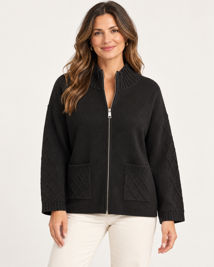 Elliott Lauren Knit Zip Up Sweater Jacket in Black, front view on model, showing a full-zip knit jacket with ribbed stand collar, silver zipper, diamond cable patch pockets, and cable-knit sleeves, styled with cream trousers