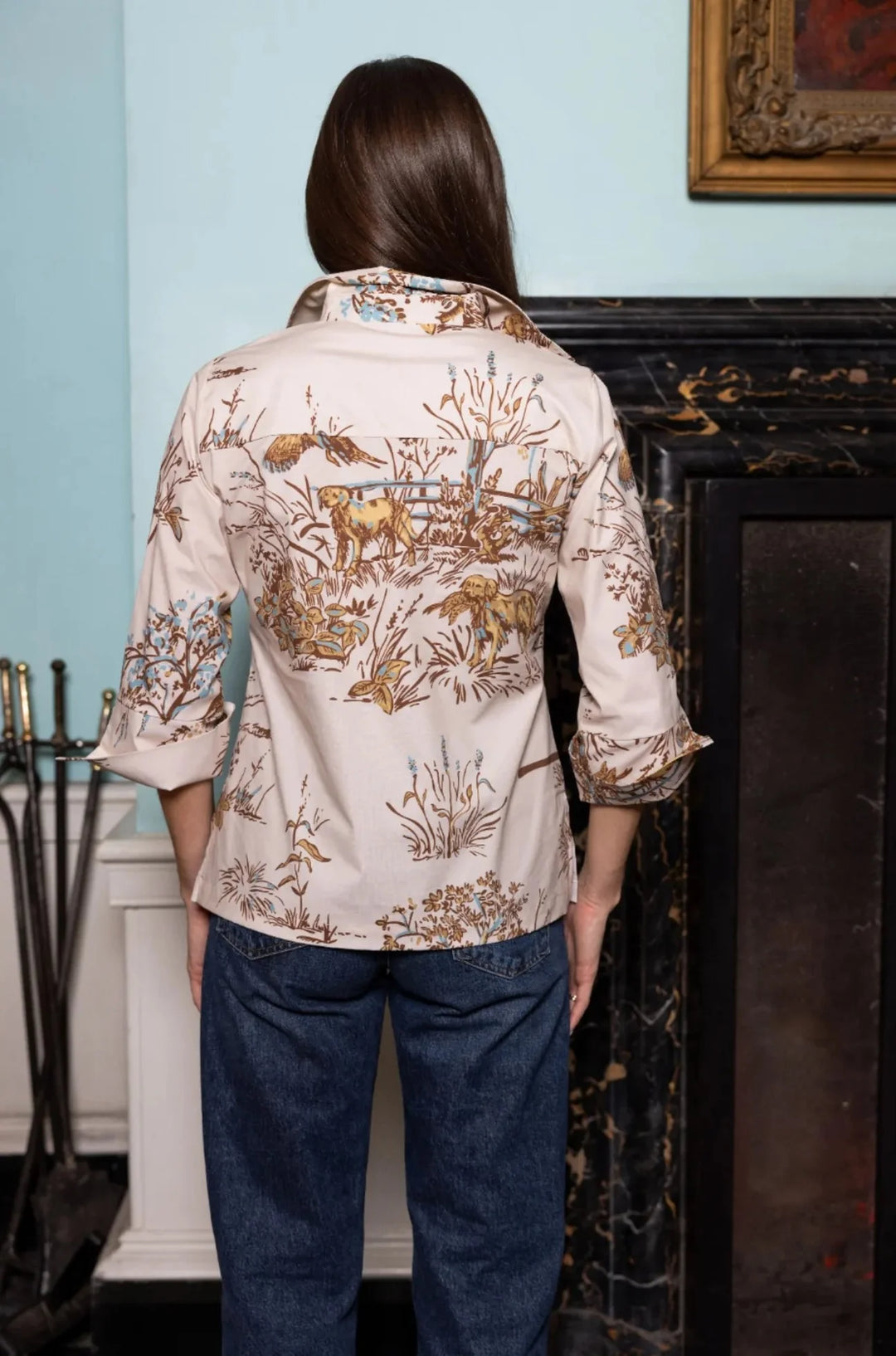 Back view, styled lifestyle: House of MBR Ruthie Top in Maple Field &amp; Feather© print shown from behind near a marble fireplace, highlighting the collar, yoke seam, split cuffs, and pastoral print across the back.