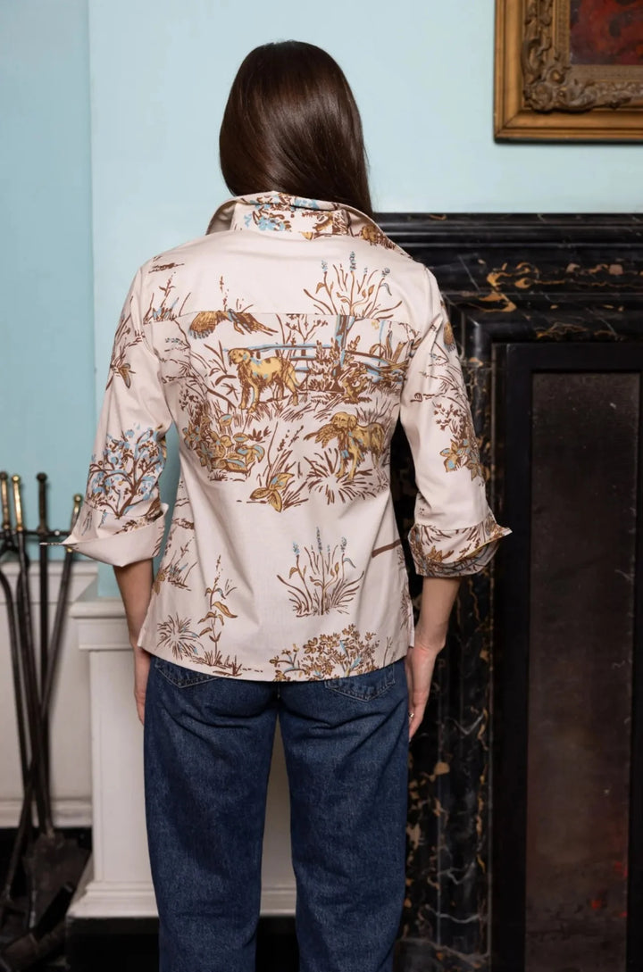 Back view, styled lifestyle: House of MBR Ruthie Top in Maple Field &amp; Feather© print shown from behind near a marble fireplace, highlighting the collar, yoke seam, split cuffs, and pastoral print across the back.