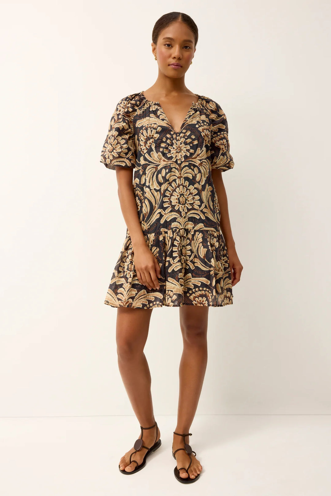Marie Oliver Greta Tropez Dress in dark navy and gold medallion print — front full-length on-model view, relaxed A-line silhouette with puff sleeves and tiered mini hem, model wearing flat leather sandals