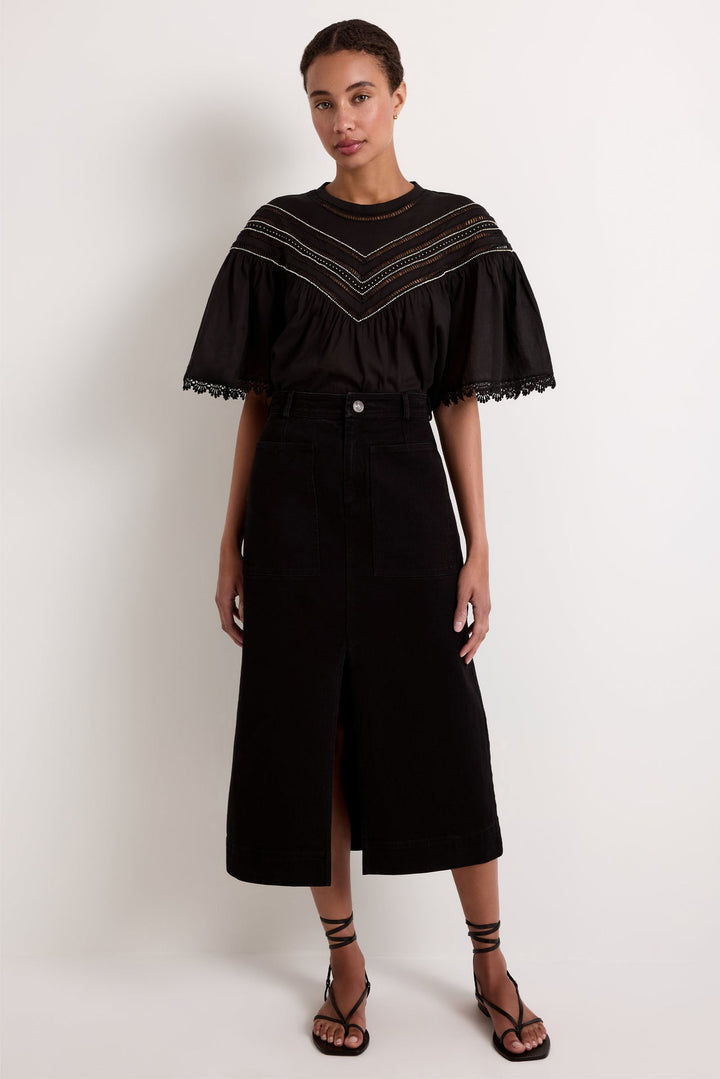Marie Oliver Leighton Denim Skirt in Pavement, full-length front view on model, showing a high-rise column midi skirt with large front patch pockets and center front slit, styled with a black embroidered flutter-sleeve blouse and black lace-up sandals
