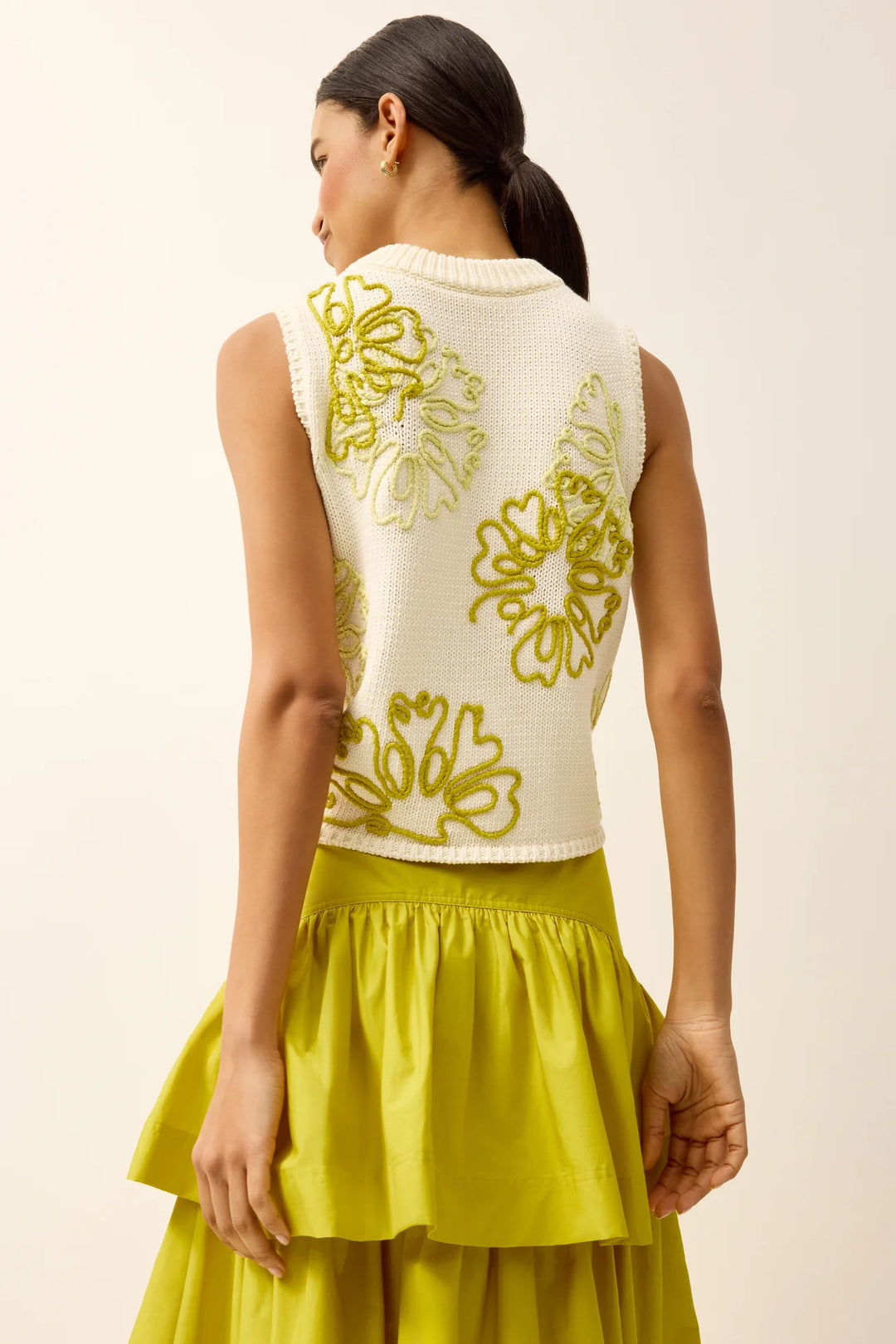 Back view of a cropped cream sleeveless knit top with oversized floral embroidery in chartreuse, worn by a model with dark hair pulled back, paired with a chartreuse tiered maxi skirt.