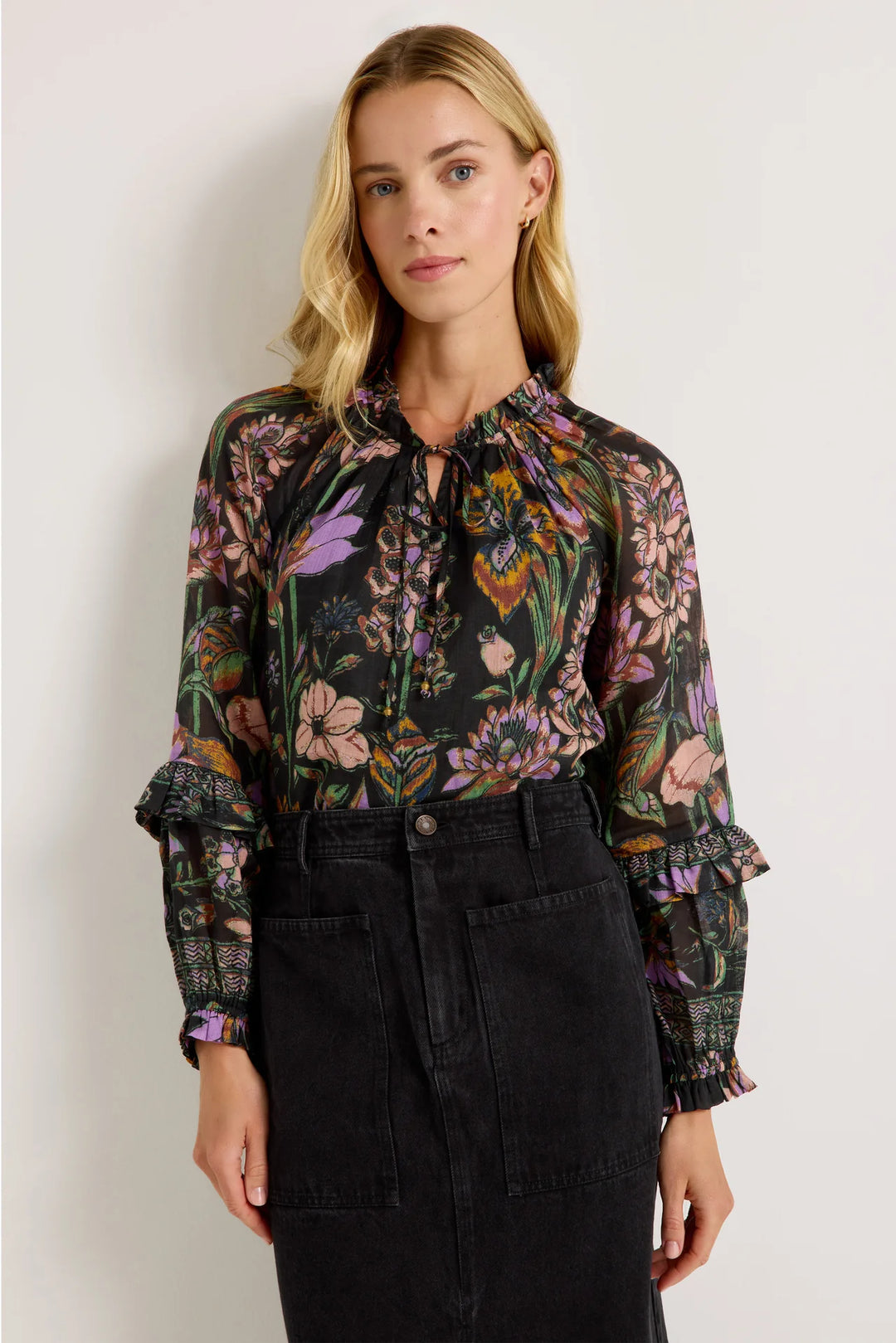 Marie Oliver Umika Top in Nightshade, front view on model, showing the ruffled neckline, bead-tipped neck ties, and violet, blush, and marigold floral print on a black ground, tucked into a charcoal denim midi skirt