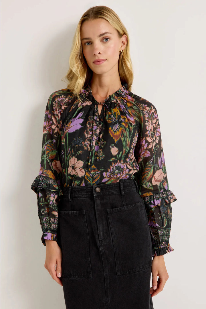 Marie Oliver Umika Top in Nightshade, front view on model, showing the ruffled neckline, bead-tipped neck ties, and violet, blush, and marigold floral print on a black ground, tucked into a charcoal denim midi skirt