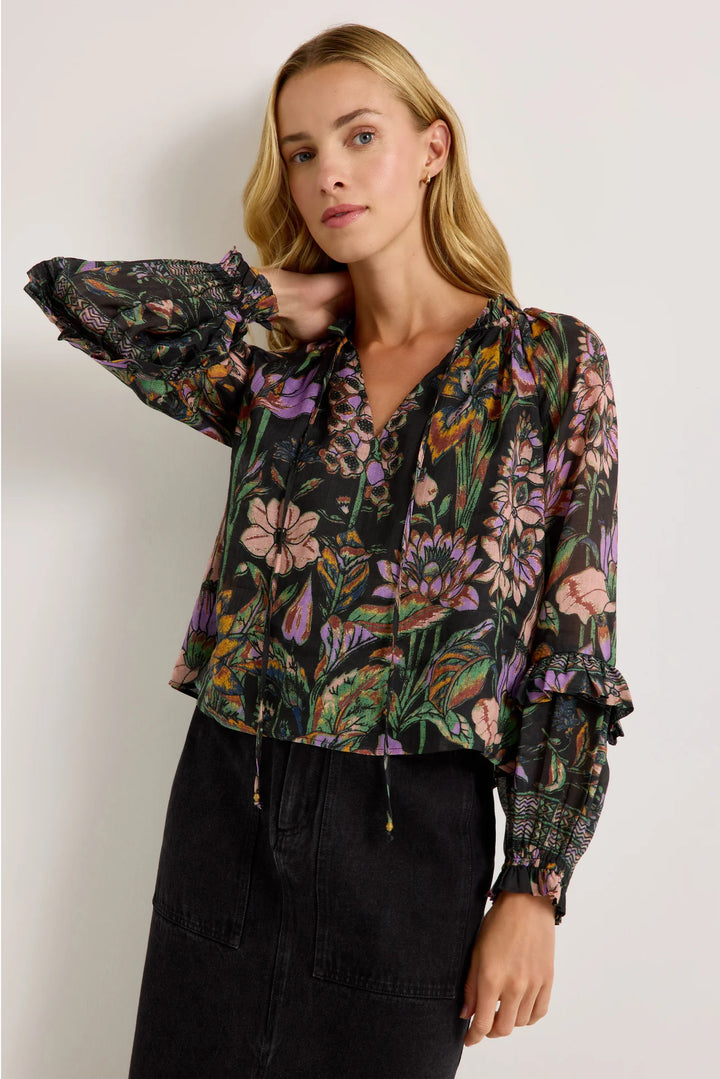 Marie Oliver Umika Top in Nightshade, three-quarter front view on model with one arm raised, showing the open V-neck, smocked ruffle cuffs, and cropped hip-length hem worn untucked