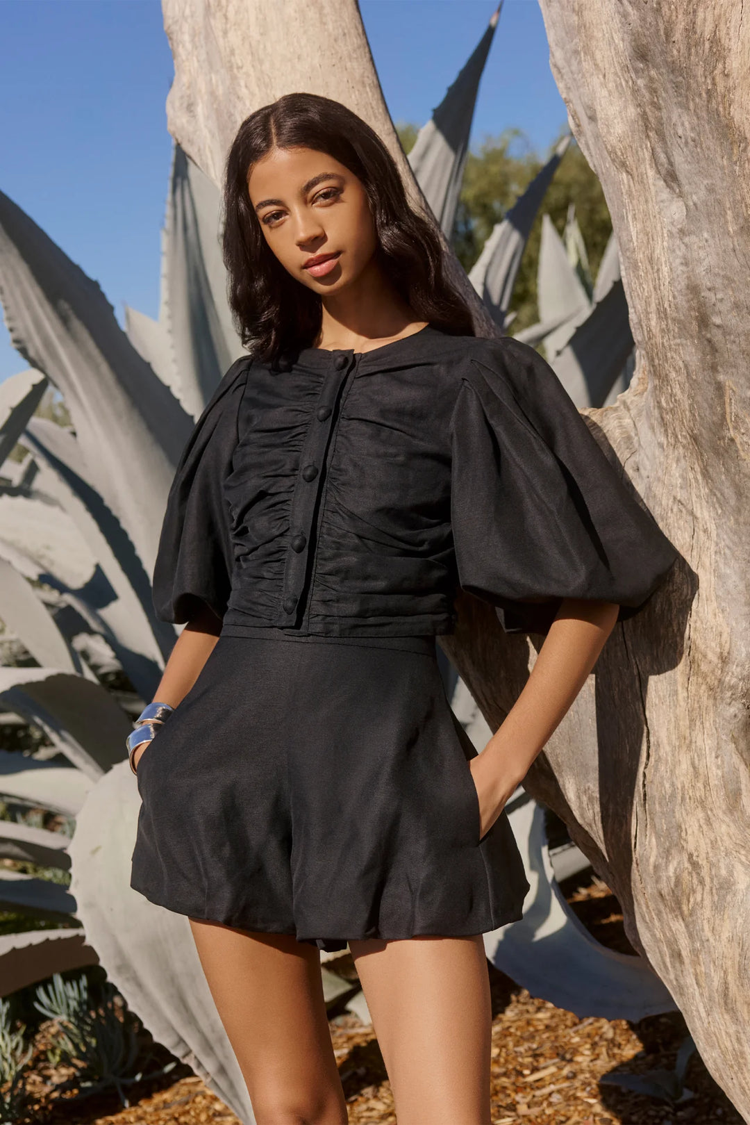 Marie Oliver Rowan Top in Black — front view showing open button closure with oversized statement buttons, ruched bodice panels, puff sleeves, and cropped fit styled with matching shorts and black tank on model outdoors