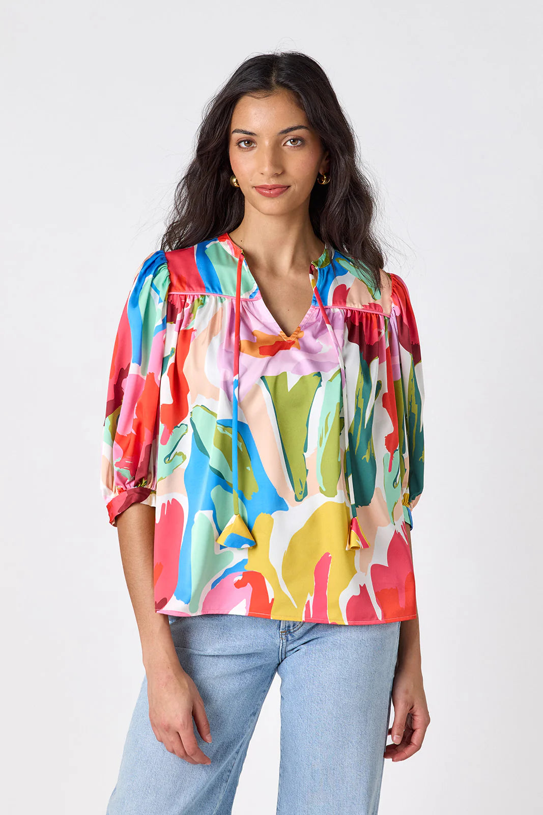 Model wearing Crosby Figgy Floral Top in Painted Petals print, cropped front view showing V-neckline with neck ties and elbow-length balloon sleeves, styled with light-wash denim and gold hoop earrings, on grey background.