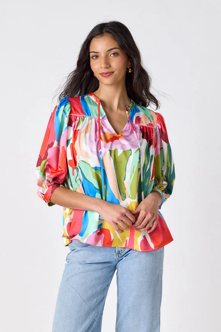 Model wearing Crosby Figgy Floral Top in Painted Petals print, cropped front view showing V-neckline with neck ties and elbow-length balloon sleeves, styled with light-wash denim and gold hoop earrings, on grey background.