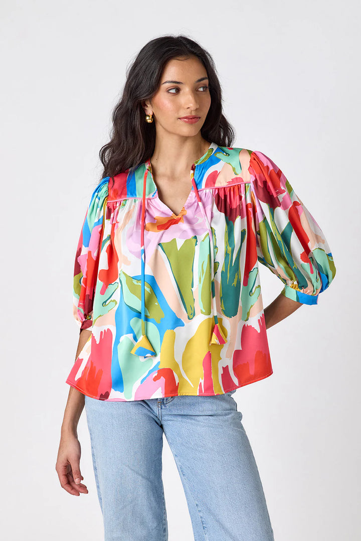Model wearing Crosby Figgy Floral Top in Painted Petals print, cropped front view showing V-neckline with neck ties and elbow-length balloon sleeves, styled with light-wash denim and gold hoop earrings, on grey background.