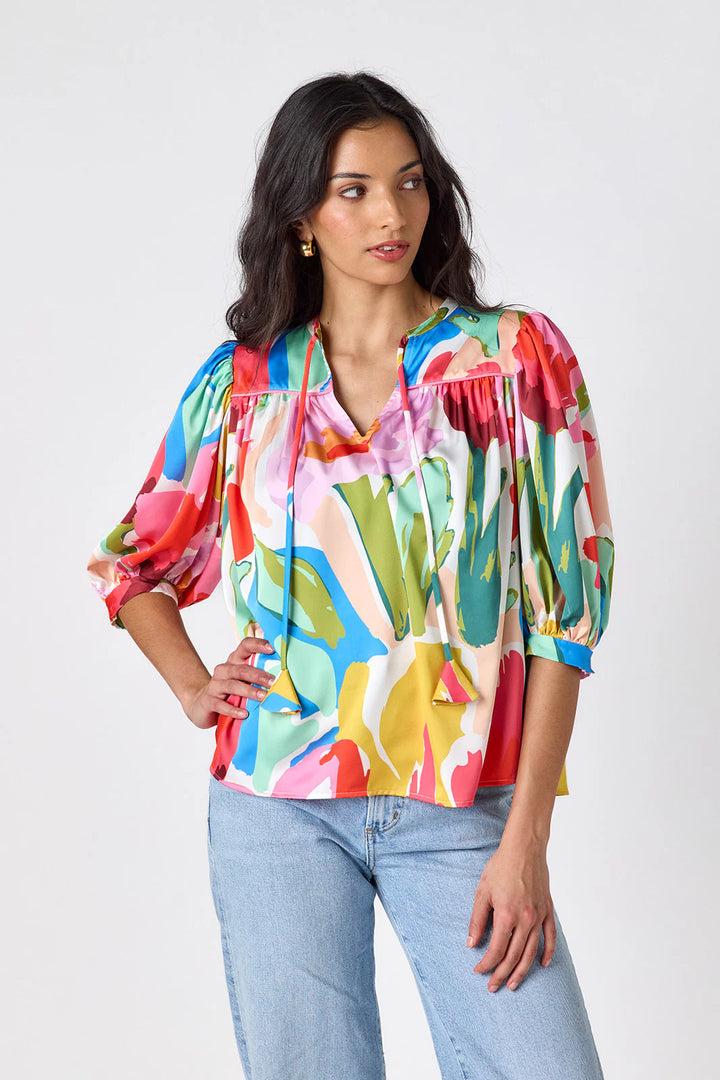 Model wearing Crosby Figgy Floral Top in Painted Petals print, cropped front view showing V-neckline with neck ties and elbow-length balloon sleeves, styled with light-wash denim and gold hoop earrings, on grey background.