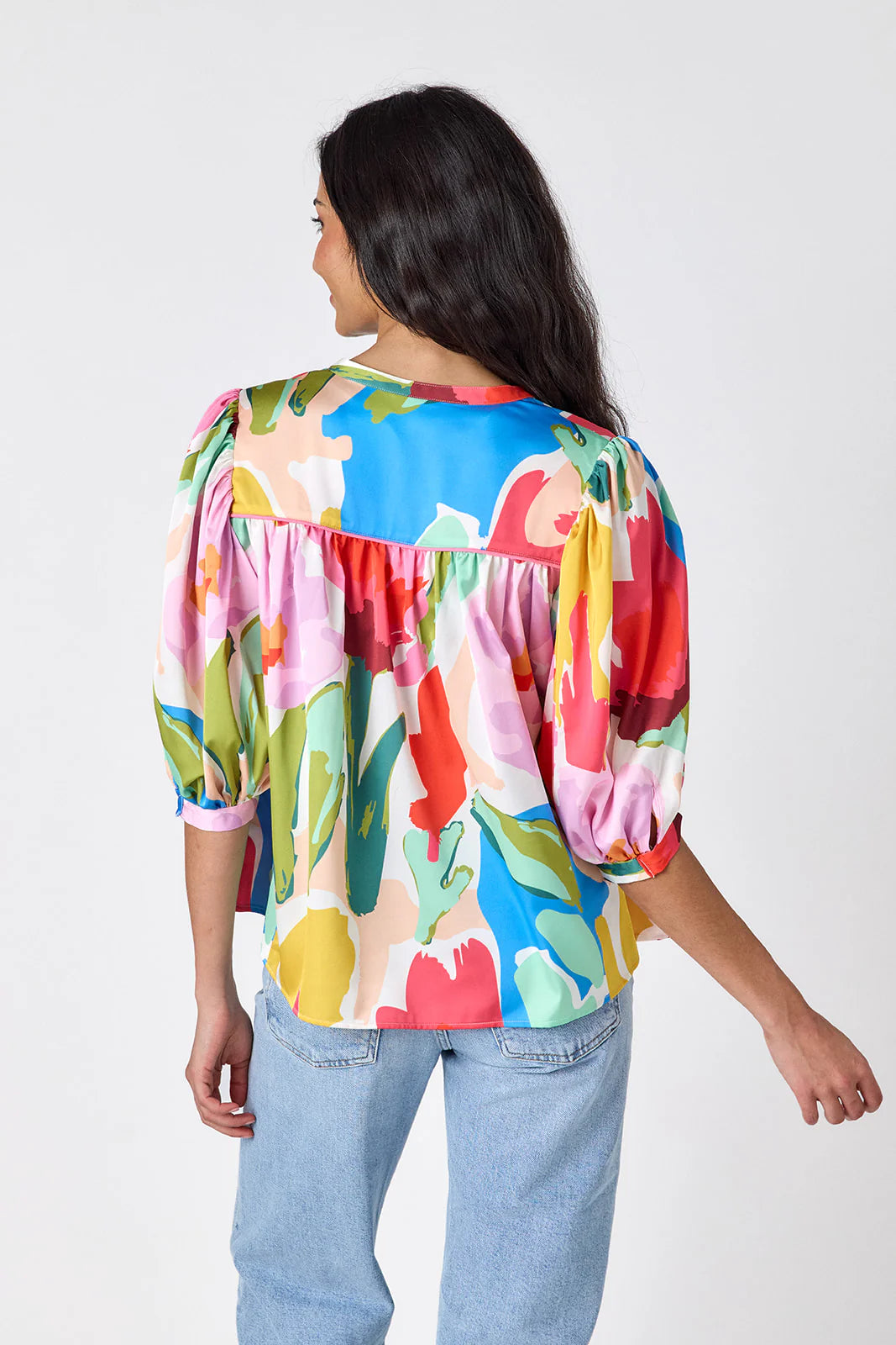Model wearing Crosby Figgy Floral Top in Painted Petals print, cropped front view showing V-neckline with neck ties and elbow-length balloon sleeves, styled with light-wash denim and gold hoop earrings, on grey background.