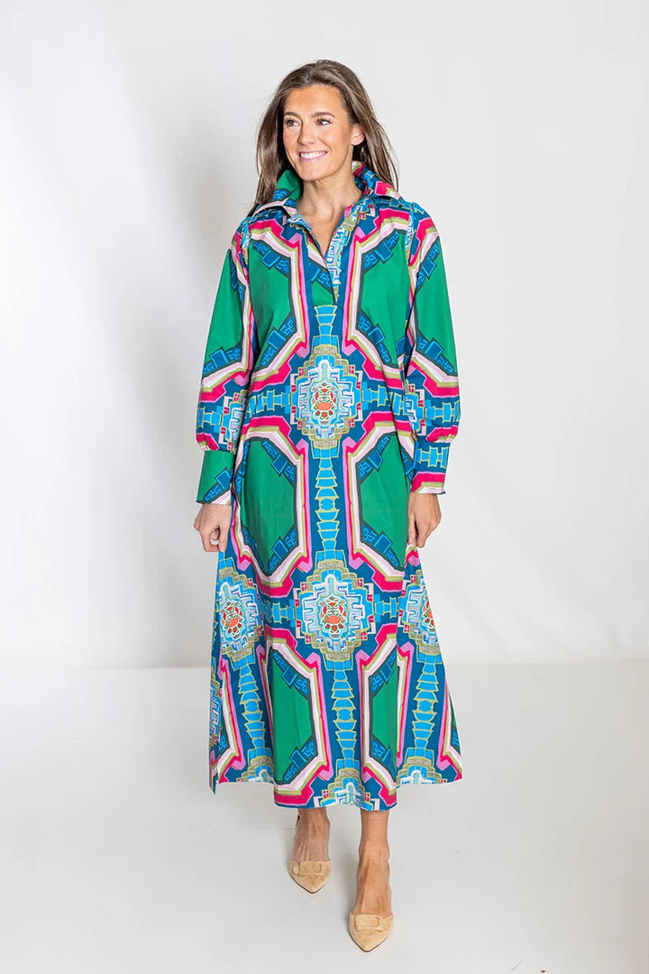 Front view, smiling: House of MBR Diana Dress Long in Portico© print, showing flowing maxi silhouette with emerald green, blue, and pink architectural medallion print, polished collar, and button-front placket, styled with nude heels.