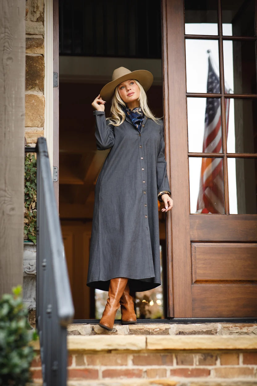 Holly Shae Adele Long Dress in Black Washed Denim, full-length front view on model standing in a wooden doorway, showing relaxed A-line maxi shape, button front, long sleeves, and mid-calf hem with cognac boots.