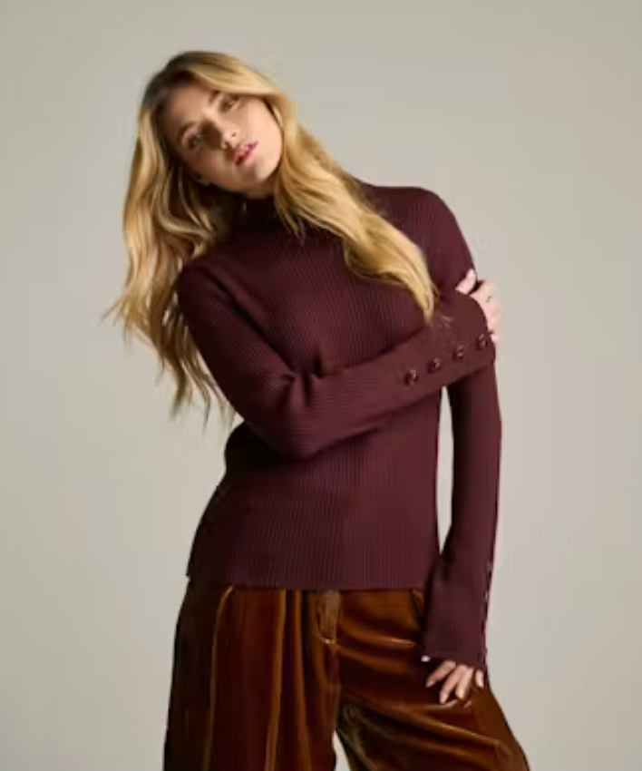 Front view of a woman wearing Rib Ready in rum raisin, a fitted ribbed mock-neck sweater with button-detail cuffs, worn with brown velvet trousers.