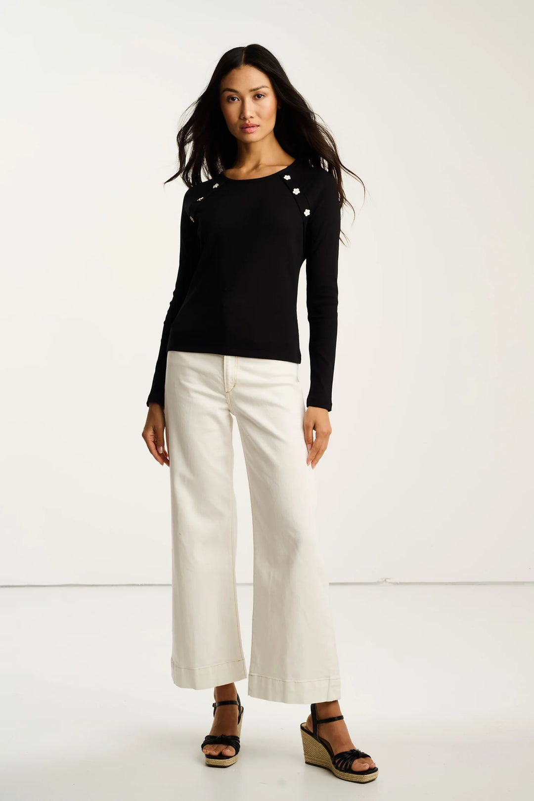 Woman wearing a black long-sleeve top with white buttons and white pants on a white background
