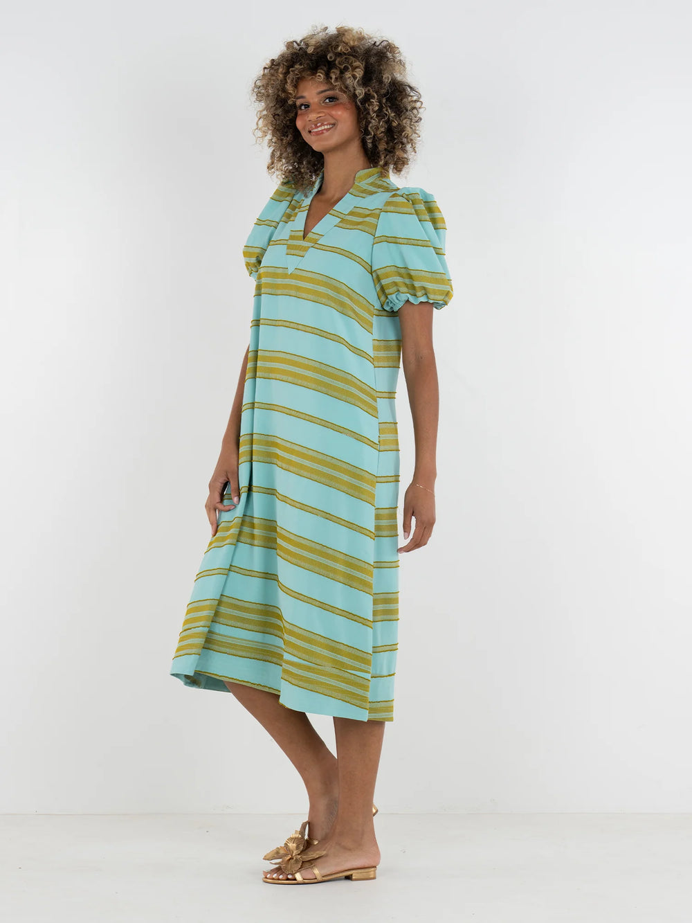Side view of Emily McCarthy Hampton Dress in Fringe Stripe, aqua and chartreuse horizontal stripe midi with V-notch mandarin collar and elastic puff sleeves, shown on model with gold sandals.