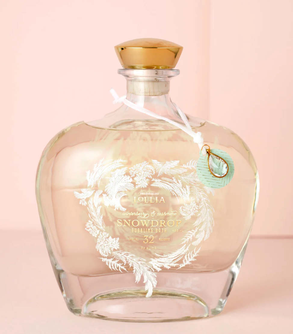 Lollia Snowdrop Limited Edition Bubbling Bath in a drop-shaped glass bottle with etched frost motif, gold cork closure, white velvet ribbon, and crystal charm against a blush background.