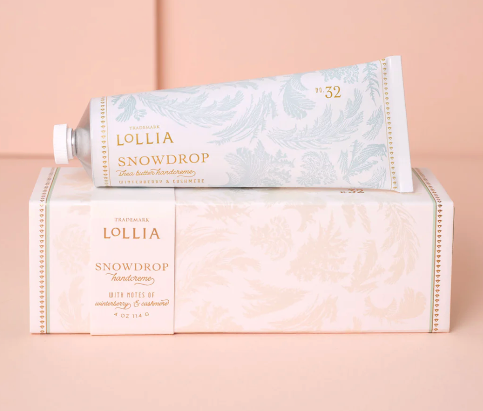 Lollia Snowdrop Shea Butter Handcreme aluminum tube resting atop its blush frost-printed gift box with gold typography, shown against a blush background.