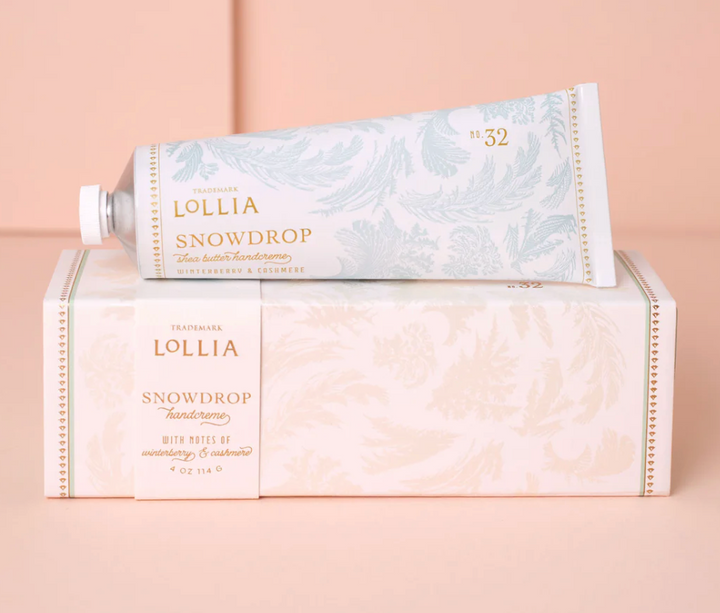 Lollia Snowdrop Shea Butter Handcreme aluminum tube resting atop its blush frost-printed gift box with gold typography, shown against a blush background.