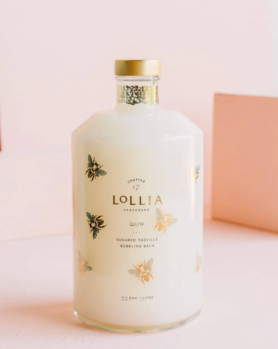 Lollia Wish Bubbling Bath in a milky white rounded glass decanter with illustrated gold and blue bees, gold cap, and decorative neck band, shown against a blush background.