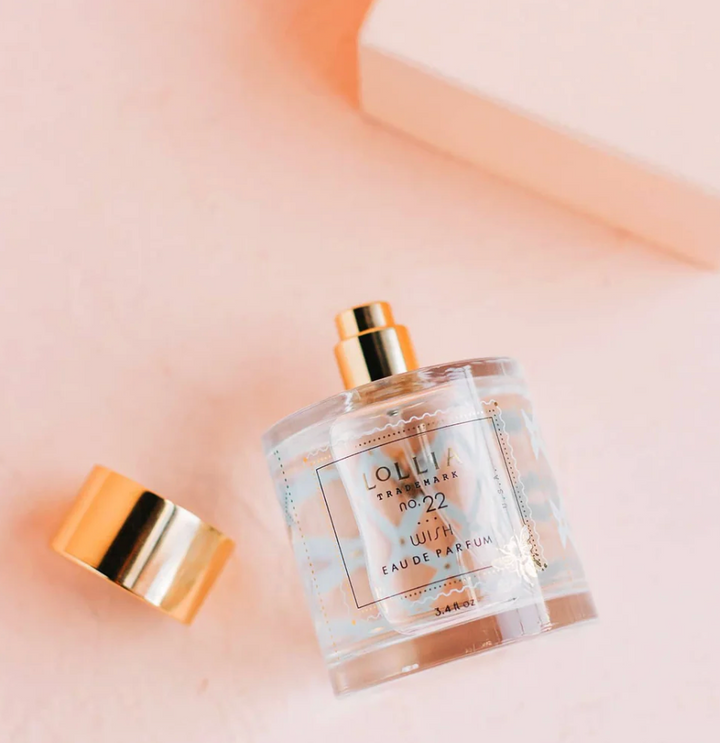 Lollia Wish Eau de Parfum bottle lying on its side with gold cap beside it, showing blue foulard pattern and stamp-style label detail.