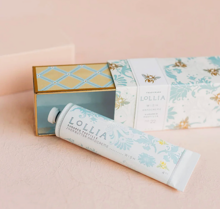 Lollia Wish Shea Butter Handcreme tube resting against its blue foulard bee-print sleeve box and open gold and blue keepsake interior tray, shown on a blush surface.