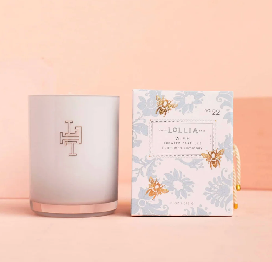 Lollia Wish Perfumed Luminary Candle in frosted white monogrammed glass vessel beside its blue damask and gold bee gift box with knotted cord, shown against a blush background.