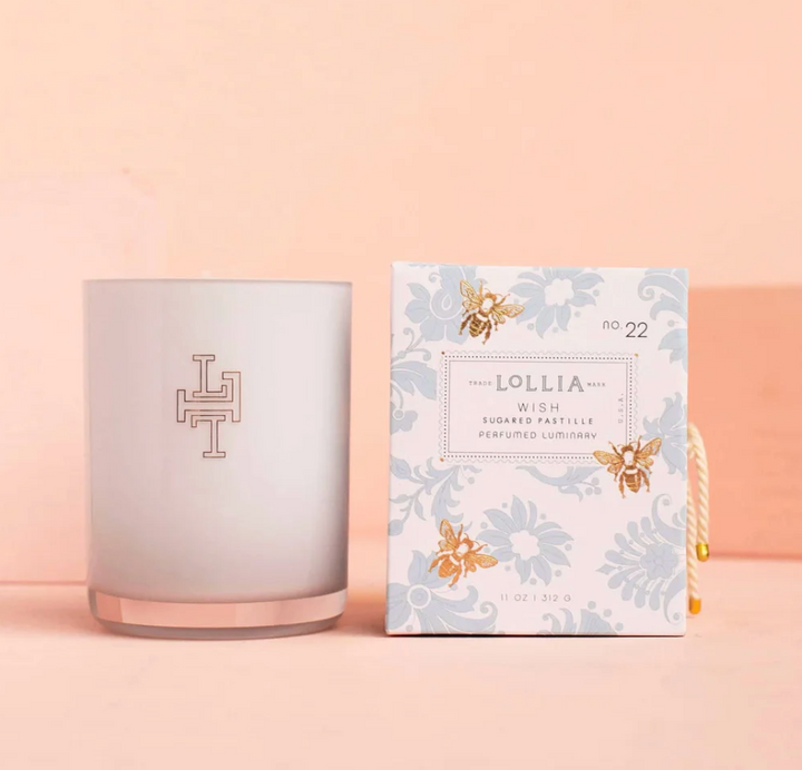 Lollia Wish Perfumed Luminary Candle in frosted white monogrammed glass vessel beside its blue damask and gold bee gift box with knotted cord, shown against a blush background.