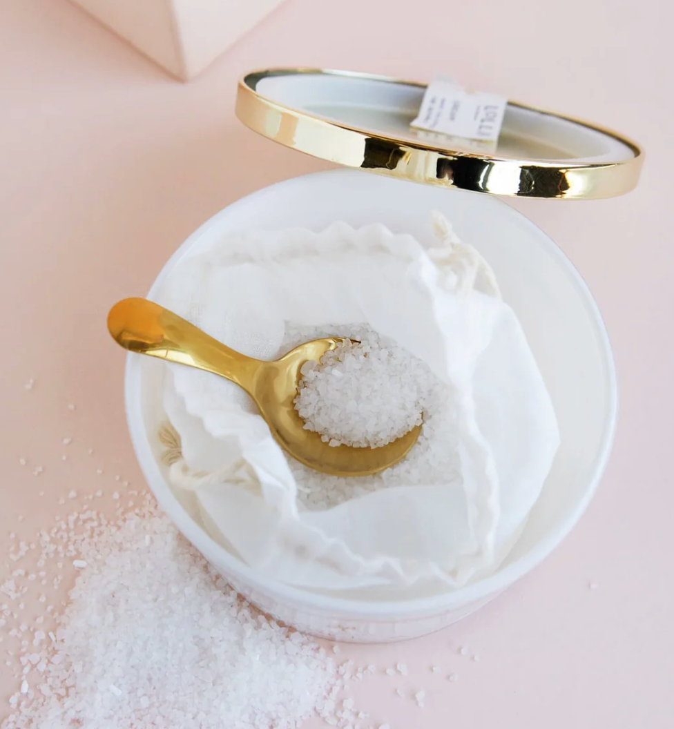 Open Lollia Wish Bath Salt jar with linen drawstring bag and gold spoon resting on coarse sea salt, lid propped open to reveal reflective interior.
