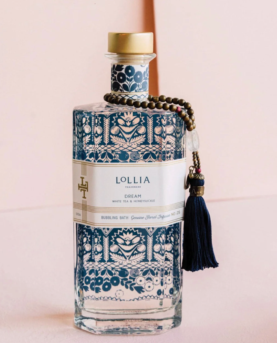 Lollia Dream Bubbling Bath in a clear glass bottle with royal blue folk-floral print, gold accents, beaded strand, navy handmade tassel, and gold cork cap, shown against a blush background.