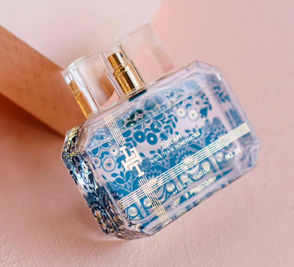 Lollia Dream Eau de Parfum in a faceted clear glass bottle with royal blue folk-floral print and gold monogram detailing, gold pump, shown at an angle against a blush background.