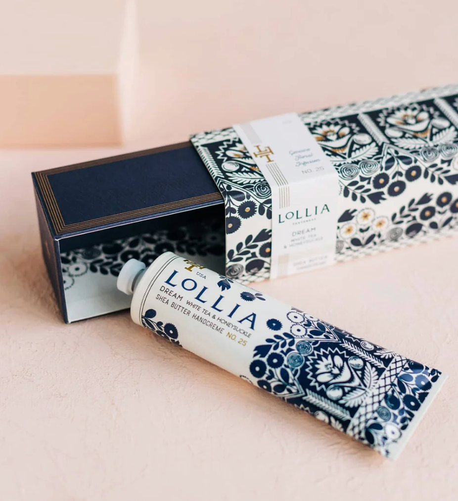 Lollia Dream Shea Butter Handcreme tube resting against its open navy keepsake box with folk-floral sleeve, shown on a blush surface.