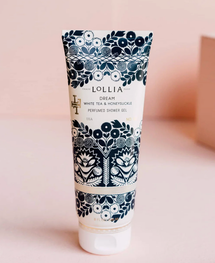  Lollia Dream Perfumed Shower Gel in a white satin-touch tube with bold navy folk-floral print and gold monogram detail, shown upright against a blush background.
