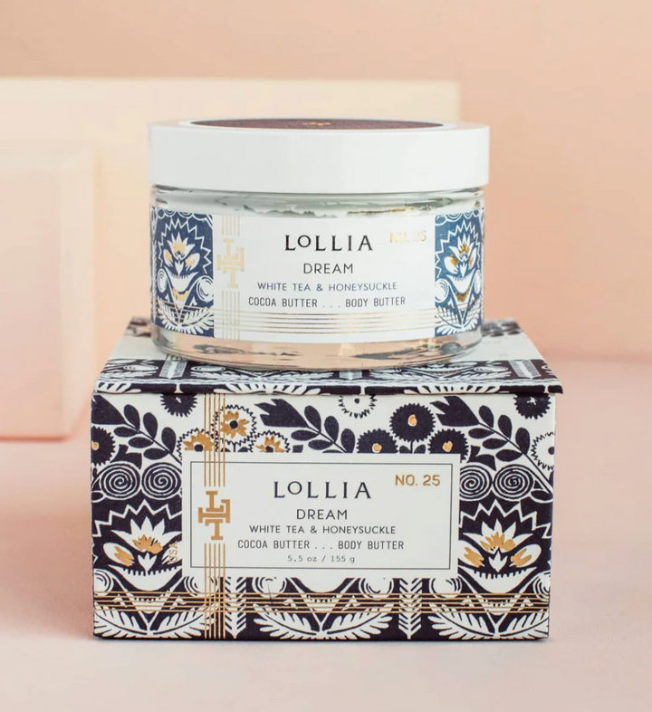 Lollia Dream Cocoa Butter Body Butter in a clear glass jar with navy folk-floral label, resting atop its navy and white gift box with gold accents, shown against a blush background.