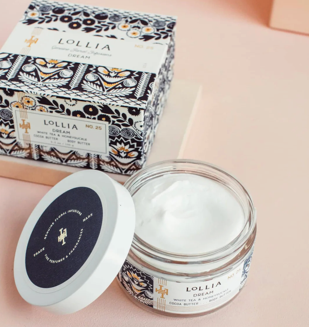 Open Lollia Dream Body Butter glass jar with white whipped texture visible and navy printed lid beside it, gift box with gold folk-floral print shown behind.