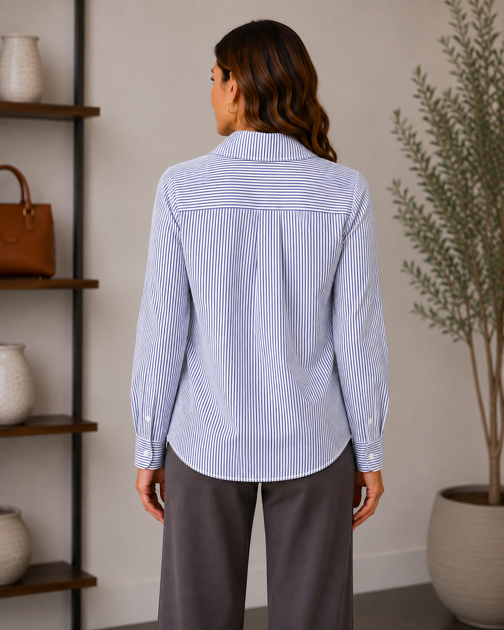 A woman shown from the back wearing a light blue and white vertically striped button-down shirt with dark trousers, standing in a styled indoor room with shelves and a potted plant.