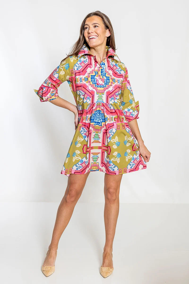 Front view, smiling: House of MBR Blythe Dress Short in Surcie© print, showing short empire-waisted silhouette with pink, olive, and blue ornamental print, shirt collar, and cuffed 3/4 sleeves, styled with nude heels.