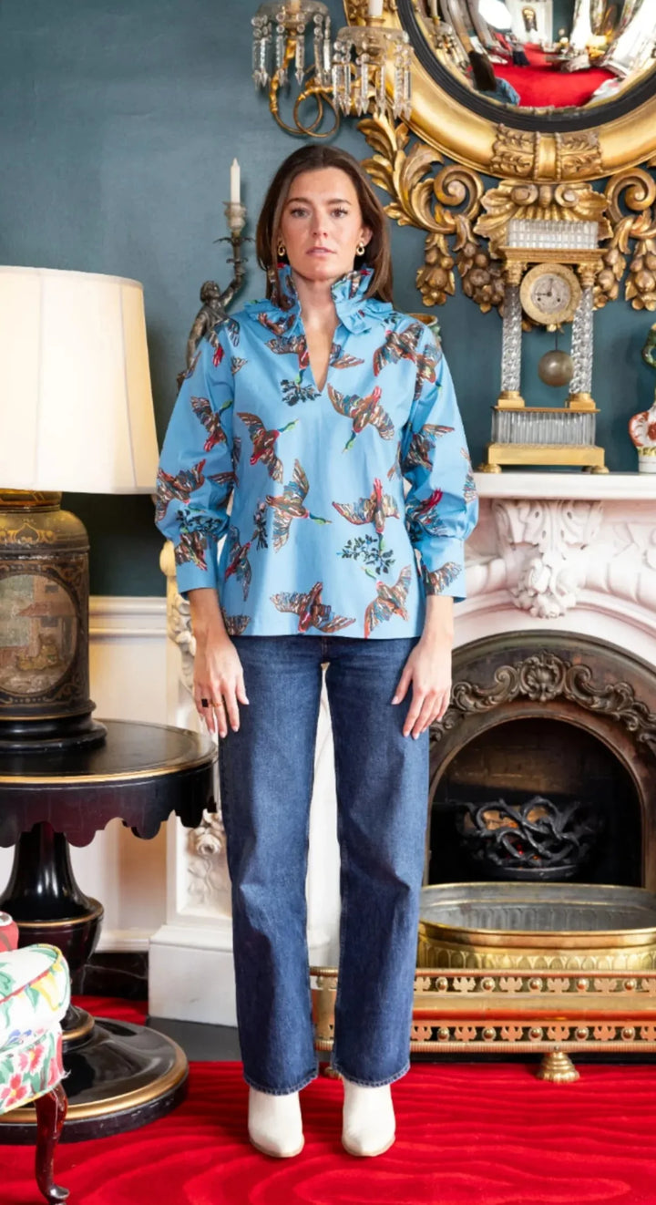 Front view, styled lifestyle: House of MBR Harper Top in Berry Grove© print, showing tunic-length silhouette in blue with illustrated bird print, double ruffle mandarin collar, and push-up sleeves, styled with dark denim in an ornate interior.