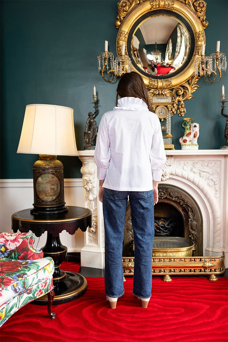 Back view, styled lifestyle: House of MBR Harper Top in Classic White shown from behind against a mantel and gilt mirror, highlighting the ruffled mandarin collar and clean silhouette, styled with dark denim.